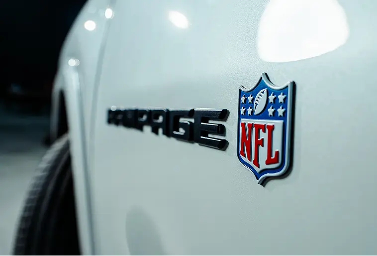 Ram: um time de campeões Foco na lateral dianteira de picape branca Ram Rampage R/T NFL Edition mostrando símbolo da NFL, que aparece nas cores originais, branco, azul e vermelho, em alto relevo. O símbolo possui formato de escudo, quatro estrelas, uma bola de futebol americano e as letras NFL. Ao lado, também em alto relevo, porém desfocado, está escrito Rampage, em preto.