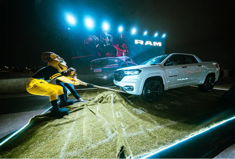 Atrações especiais RAM + NFL Mascote Ramirito, que é um carneiro montanhês estilizado, aparece auxiliando uma mulher a puxar uma Rampage por meio de uma corda que está presa no reboque dianteiro do carro, que está de lado na imagem. No chão, um tapete estilizado representa a grama com as jardas marcadas típicas do futebol americano. Ao fundo, luzes superiores iluminam a cena e é possível ver uma grande placa de anúncio da Ram.