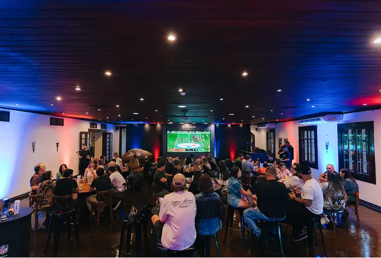 Watch Party RAM Society + NFL Ambiente escuro com luzes próximas ao teto nas cores azul e vermelho. Diversas pessoas estão no ambiente, que tem tom festivo e possui enorme telão com a transmissão de um jogo de futebol americano.
